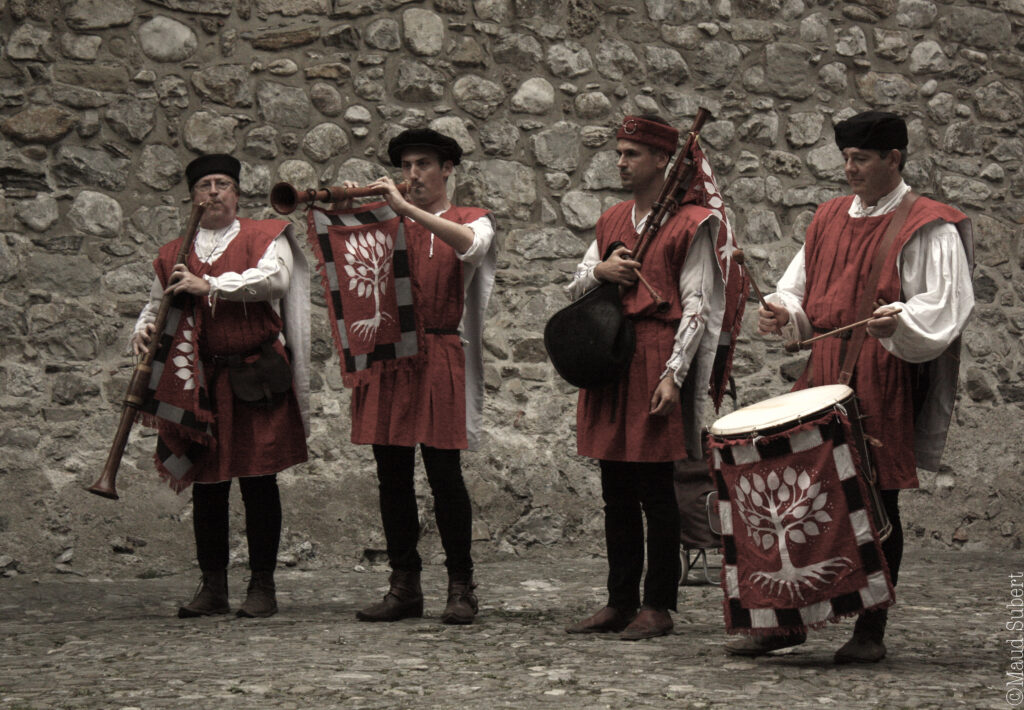 Quatre musiciens en costumes rouges jouent des instruments à vent, une cornemuse et un tambour devant un mur de pierre.