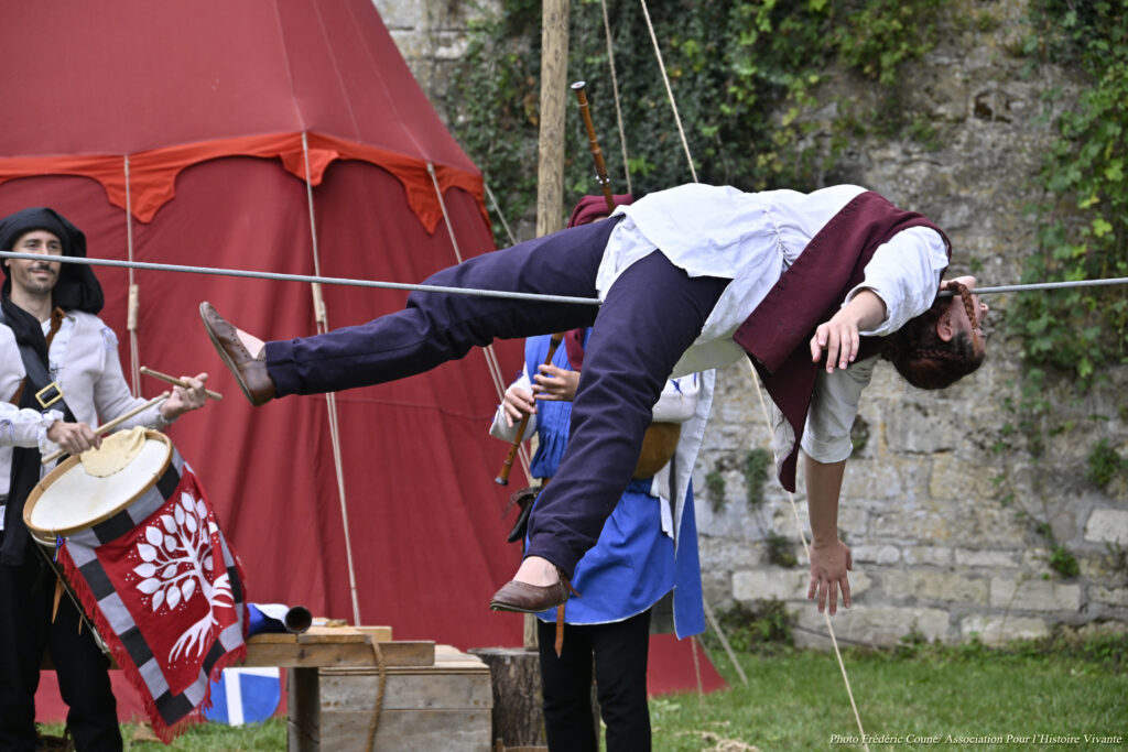 Une acrobate réalise une figure allongée sur un fil, accompagnée de musiciens en costumes historiques.